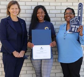Three women stand in front of a white brick wall. The woman in the middle holds a certificate and the woman on the right has a large #1 foam finger.