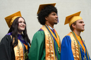 Three students pose in full graduation regalia with gold caps, stoles, and various colored cords.