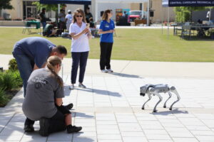 A quadruped robot walks on a cement area in the Green Space while people observe and interact nearby during a tech demonstration.