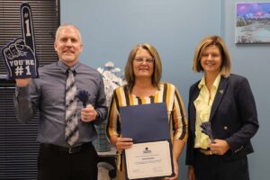 One man and two woman pose for the camera. All dressed in business casual clothes. The woman in the middle holds a certificate and the man on the right has a #1 foam finger.
