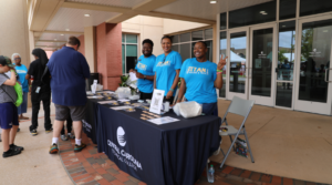 Three young adults in light blue matching tee shirts pose for the camera at a registration table outside of a college building.