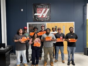 A group of young, diverse students pose with machining tool kits in front of a sign for the Gene Haas Foundation.